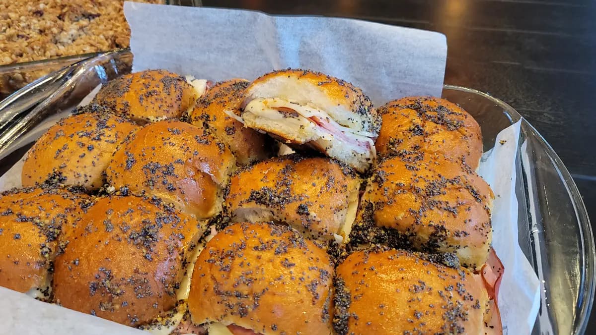 Golden-brown cheesy ham sliders on a white plate with melted Swiss cheese and poppy seeds, steam rising from the warm rolls