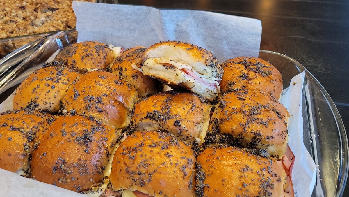 Golden-brown cheesy ham sliders on a white plate with melted Swiss cheese and poppy seeds, steam rising from the warm rolls