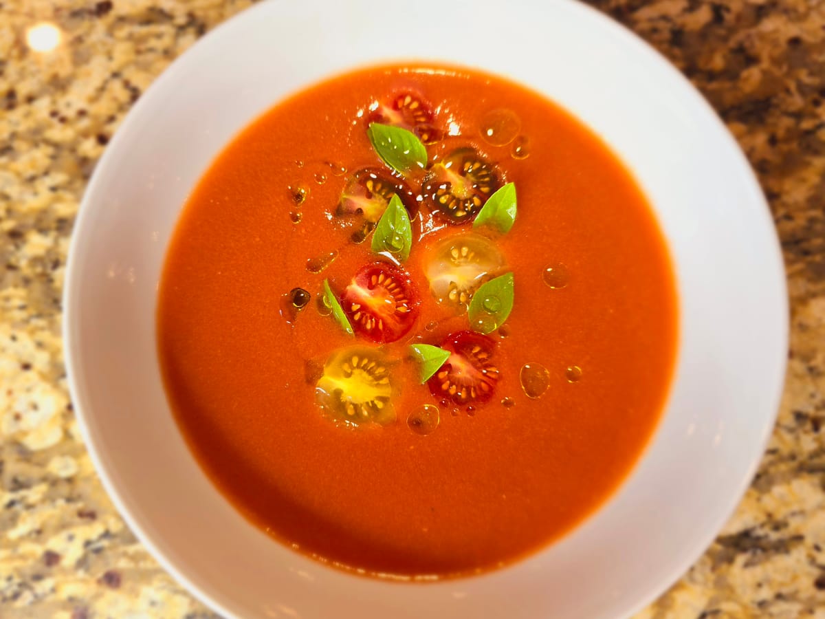 Bowl of vibrant red gazpacho soup garnished with diced vegetables and olive oil drizzle, fresh tomatoes and cucumber visible