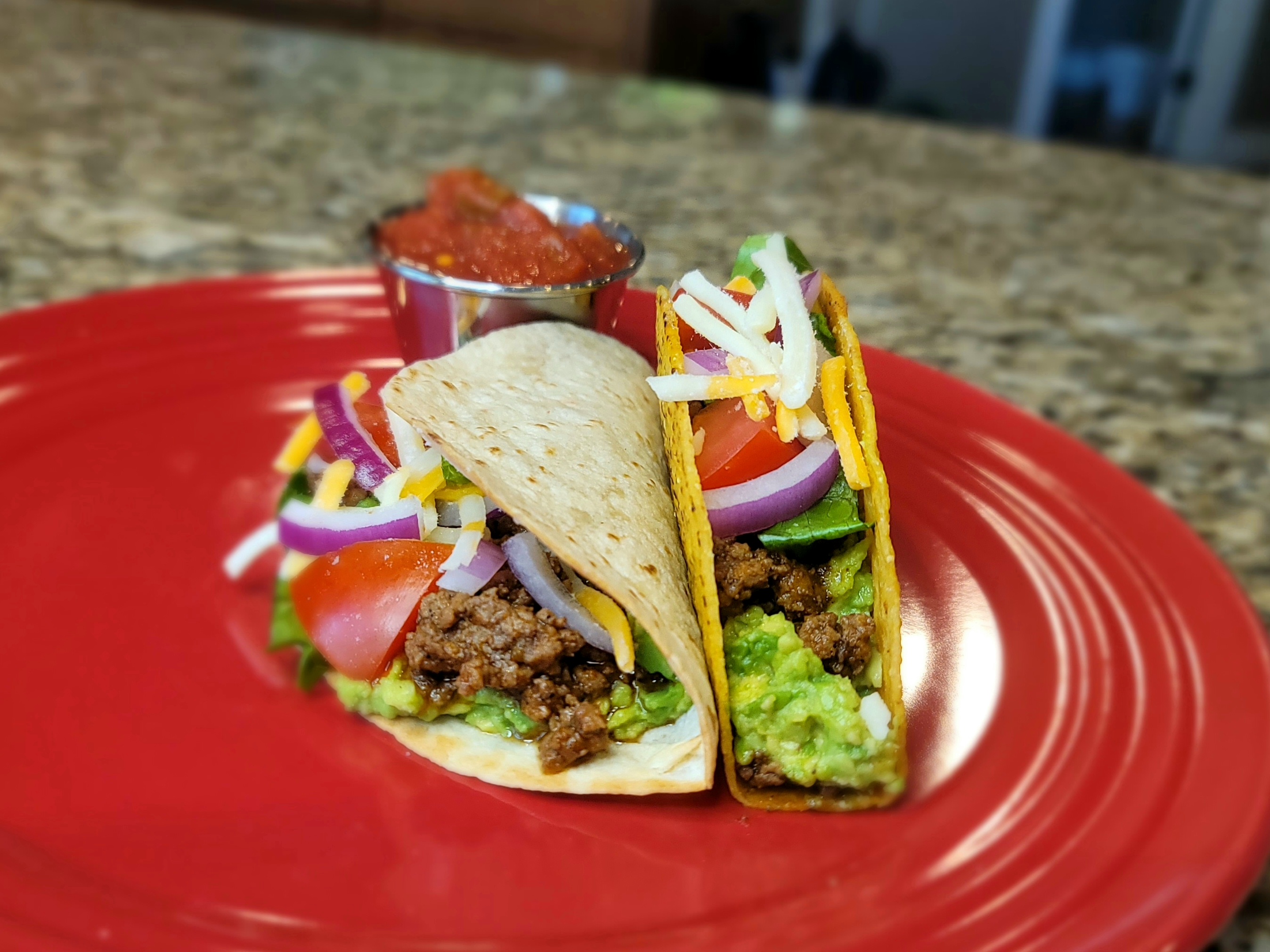 Ground beef tacos topped with lettuce, tomatoes, cheese, avocado, and salsa on a white plate