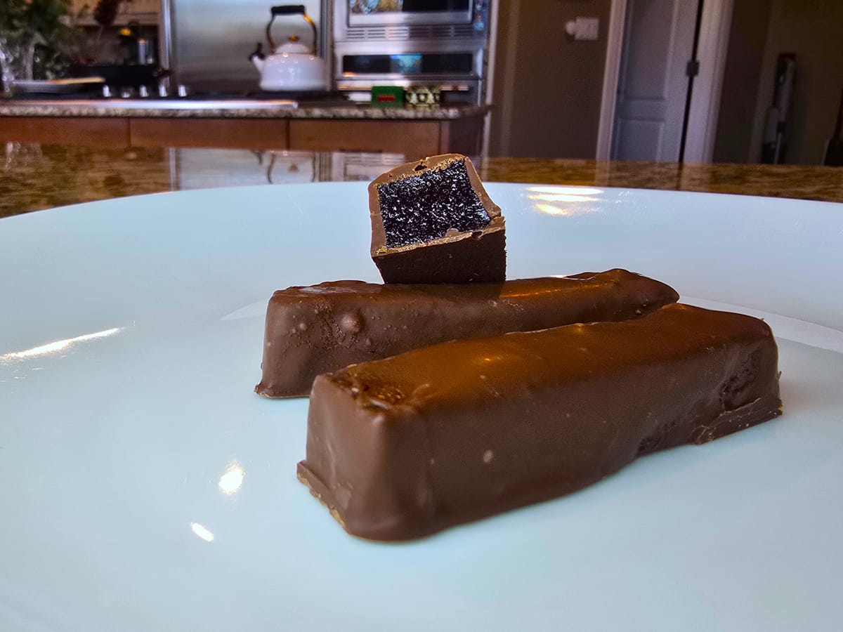 Dark chocolate-dipped raspberry jellie sticks dusted with sugar on a white surface, showing glossy chocolate coating and jewel-like raspberry centers