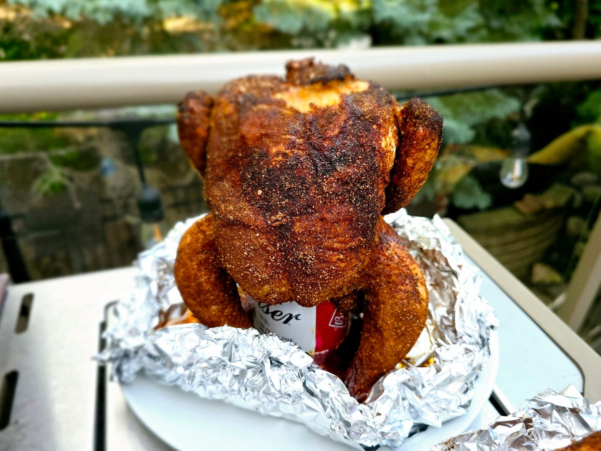 Golden-brown beer can chicken sitting upright on a grill grate with crispy, bronzed skin and fresh herb garnish