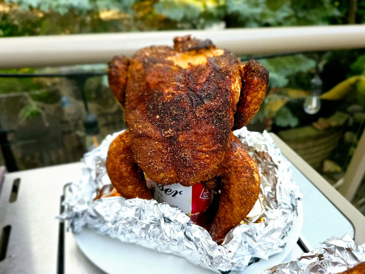 Golden-brown beer can chicken sitting upright on a grill grate with crispy, bronzed skin and fresh herb garnish