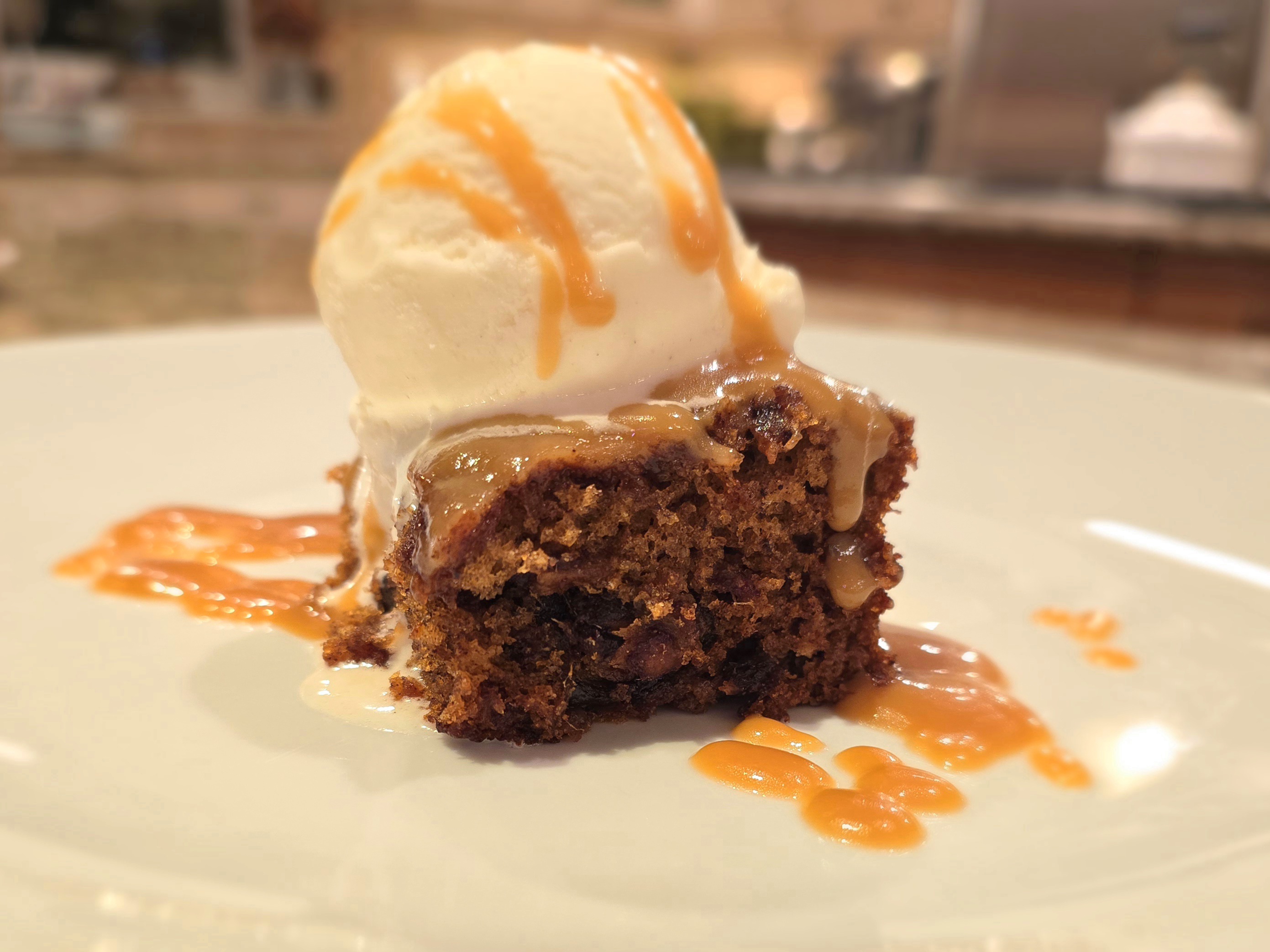 Warm sticky toffee pudding with glossy toffee sauce dripping down the sides, served with vanilla ice cream on a white plate